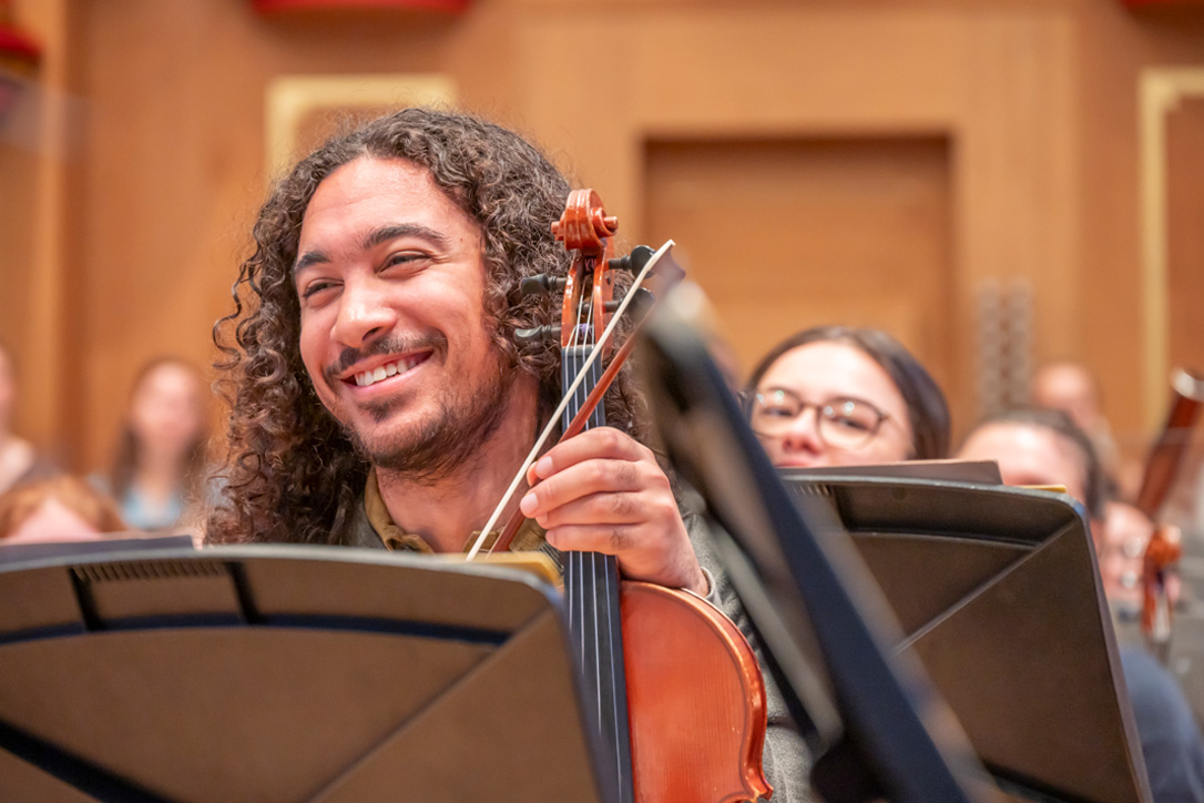 A male student, holding his violin, smiling and chatting during an orchestral rehearsal break.
